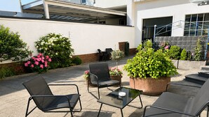 En udendørs terrasse med metalstole og et bord, omgivet af krukker med planter og blomster.