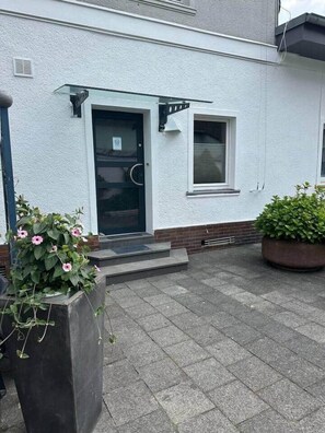 Un bâtiment blanc avec une porte noire, une petite fenêtre et un pot de fleurs contenant une plante aux fleurs roses.