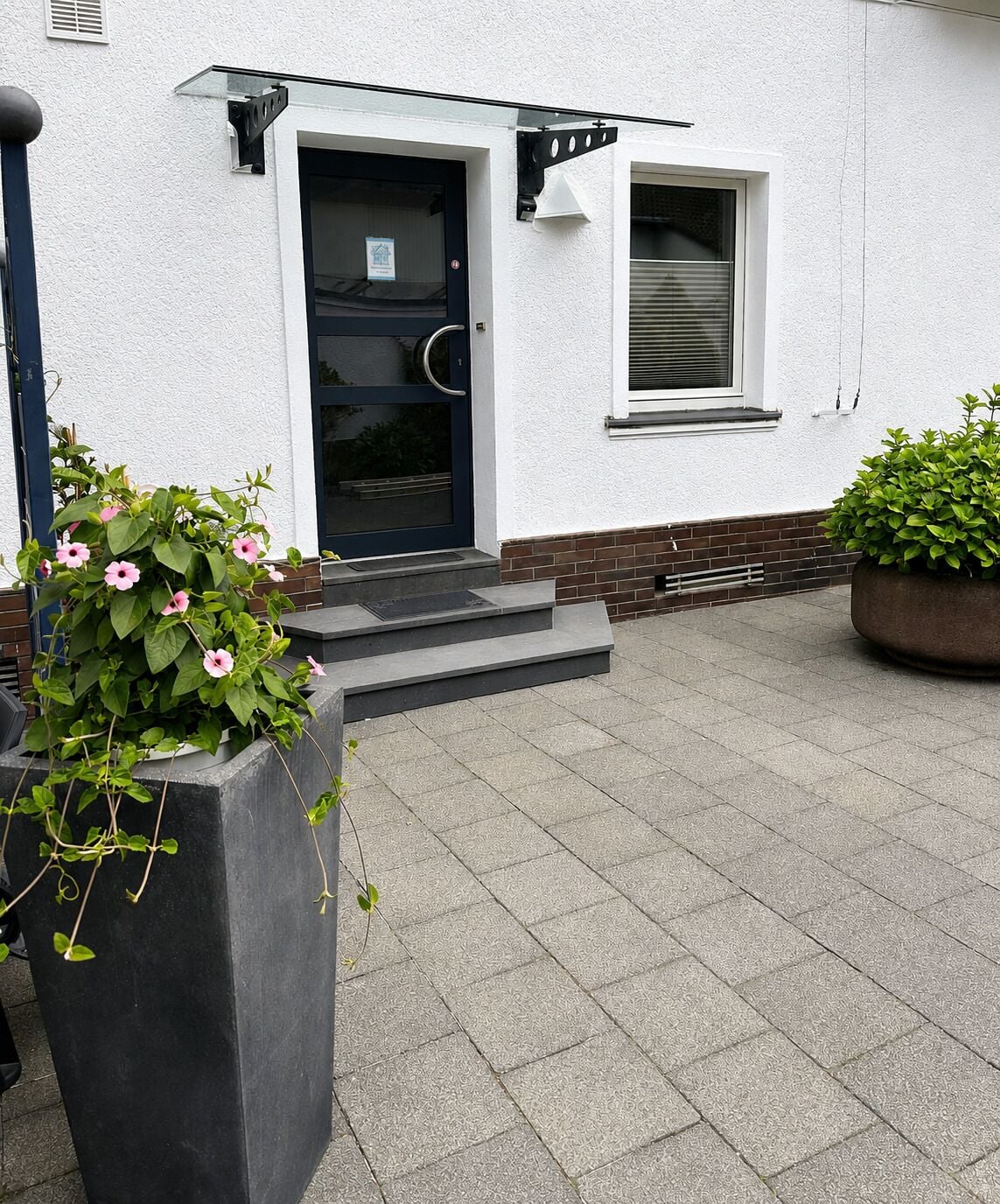 Ein Gebäude mit einer schwarzen Tür, einem kleinen Fenster und einer Ziegelbasis. Vor dem Gebäude befinden sich zwei Pflanzkästen mit Pflanzen.