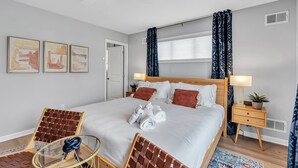 Une chambre d’hôtel moderne avec un grand lit, une table de chevet avec une lampe, une chaise et une petite table avec une plante.