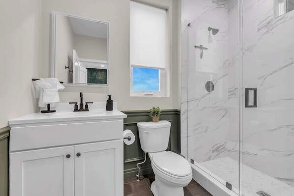 A bathroom with a white vanity, a toilet, a shower with glass walls, and a window with blinds.