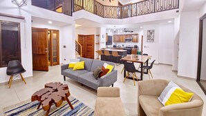 Amplia sala de estar con un sofá gris, sillones color beige y una mesa de centro. Se ve una cocina al fondo con armarios de madera y un comedor.