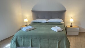 Una cama bien tendida con una colcha verde, dos almohadas blancas y dos lámparas de noche sobre mesitas de madera.