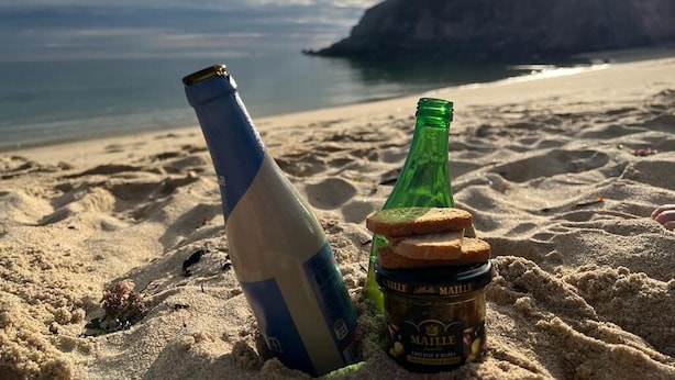 En flaske øl og et glas Maille på en sandstrand med udsigt til havet og klipper.