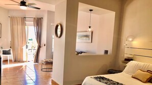 A bedroom with a bed, a bedside table, a hanging light, a wall-mounted mirror, and a door leading to another room.