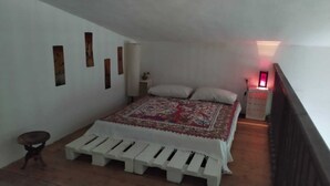 Une chambre à coucher comprenant un lit, un petit tabouret, une table de chevet et une étagère fixée au mur.