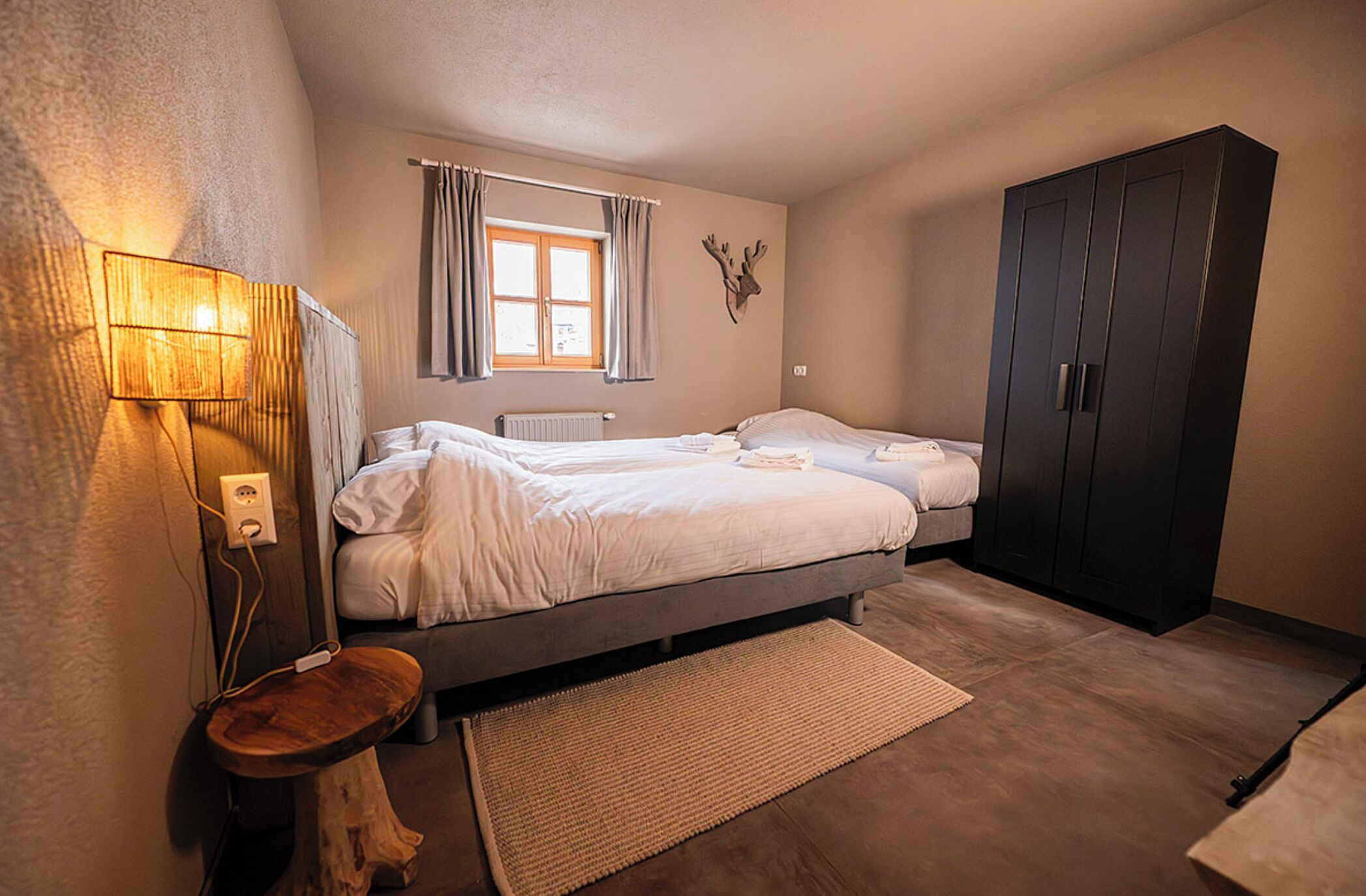 A bedroom with a bed, a wardrobe, a lamp, a window with curtains, and a wall-mounted deer head decoration.