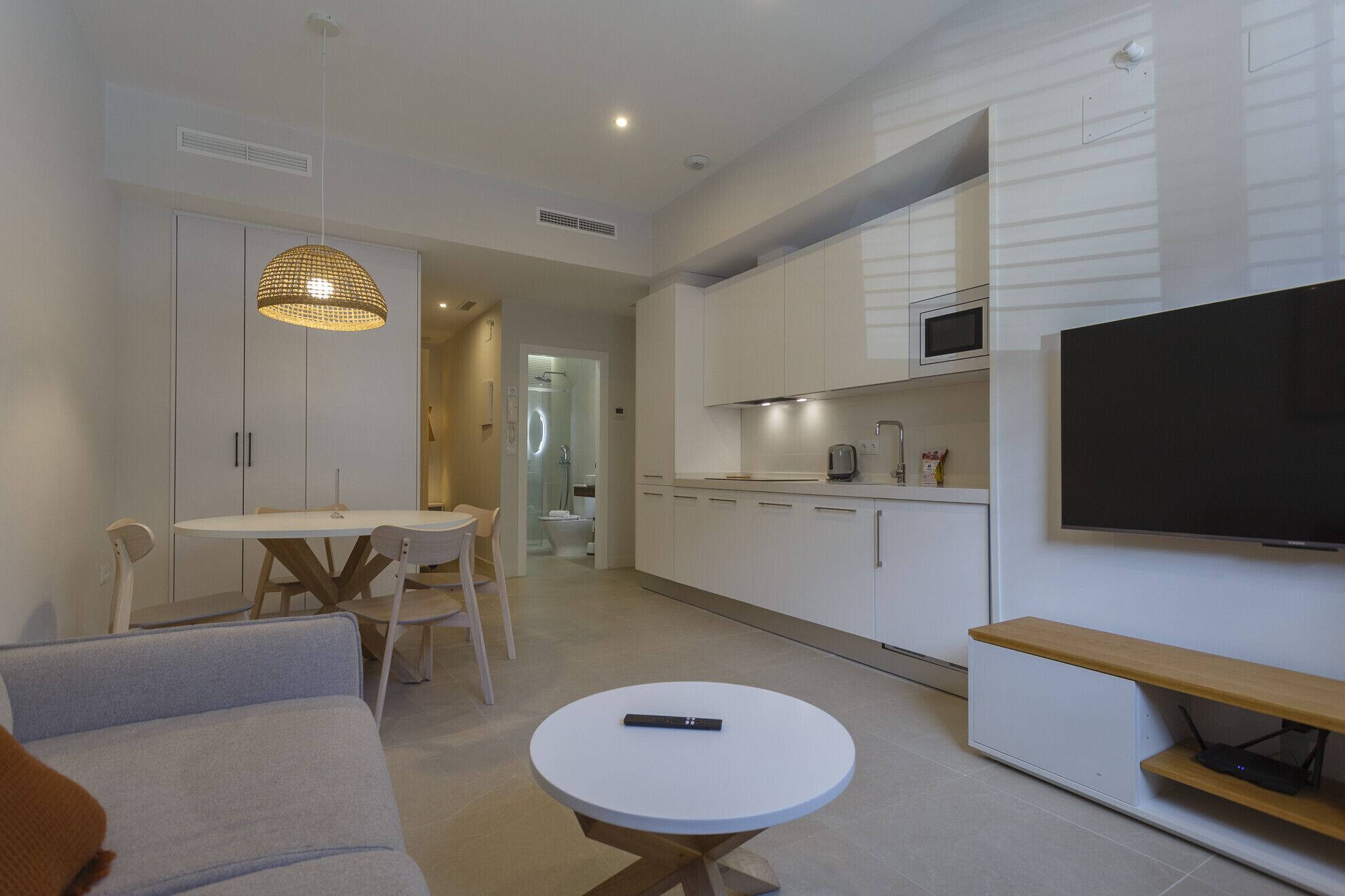 Una sala de estar moderna con un sofá, una mesa de comedor y sillas. Hay un televisor montado en la pared y una mesita con un control remoto.