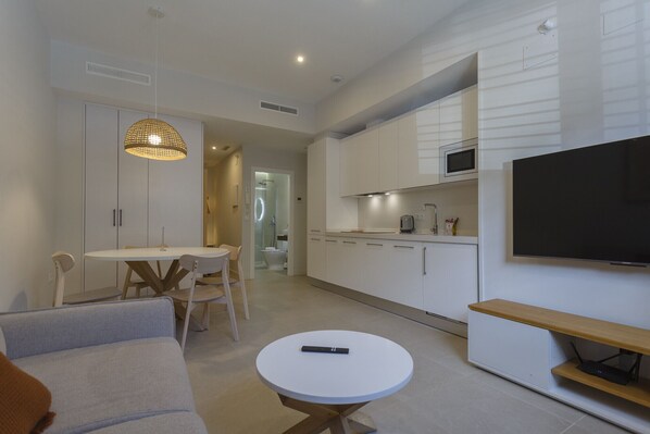 Una sala de estar moderna con un sofá, una mesa de comedor y sillas. Hay un televisor montado en la pared y una mesita con un control remoto.