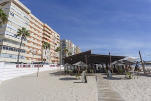 Un hotel frente a la playa con mesas al aire libre, palmeras y vista al mar.