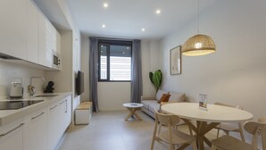 A modern kitchen with white cabinets, a dining area with a round table and chairs, a sofa, and a large window with curtains.