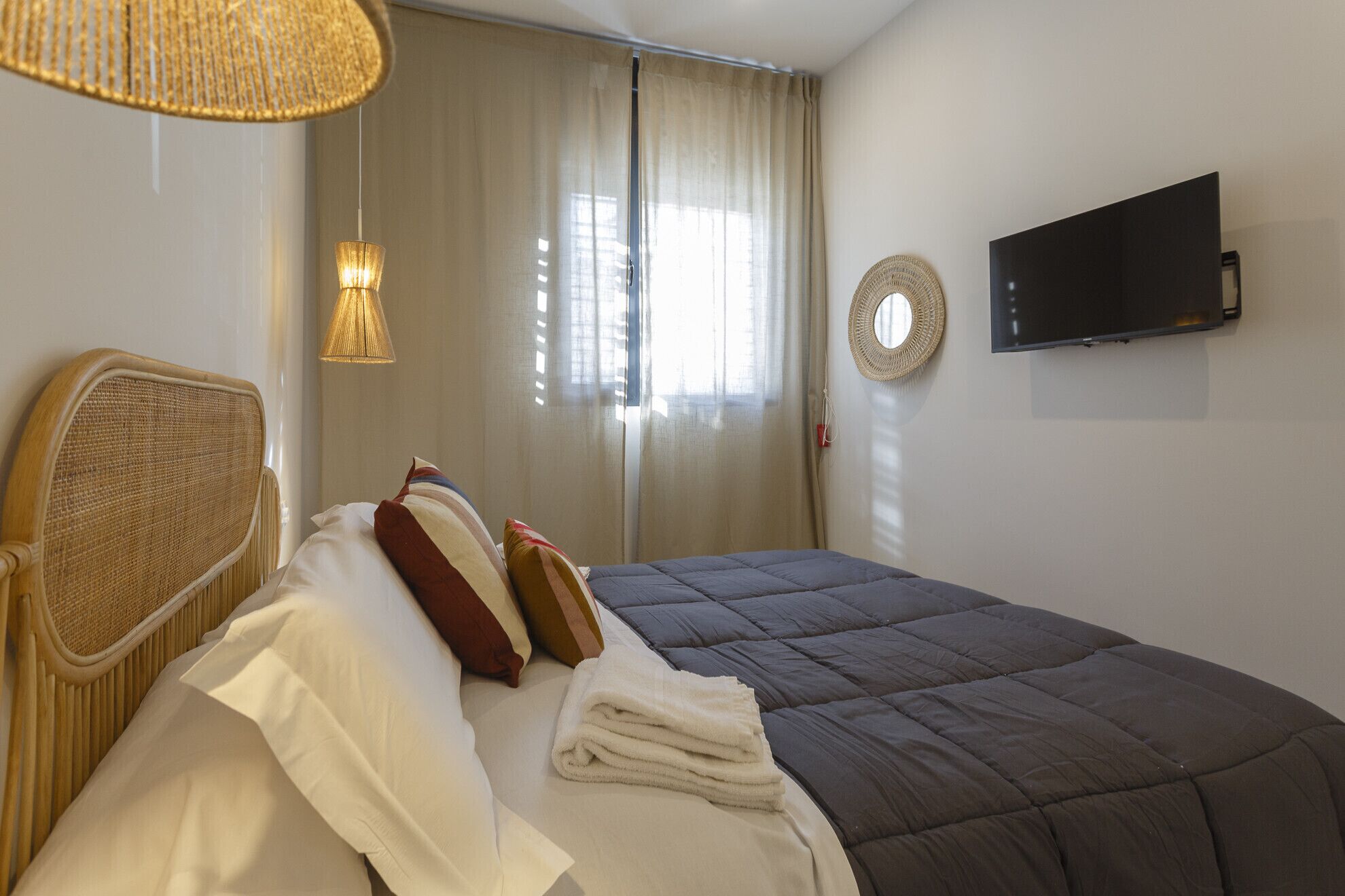 Ein ordentlich bezogenes Bett mit dunkler Tagesdecke, ein Korbstuhl, ein Wandbehang aus Gewebe und ein an der Wand befestigter Flachbildfernseher.