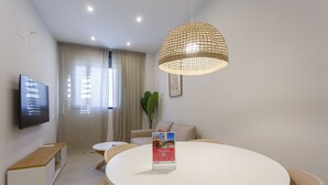 Et moderne spiseområde med et rundt hvidt bord, træstole, en hængende vævet lampe, et fladskærms-tv monteret på væggen og en potteplante.