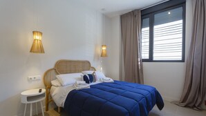 A bedroom with a bed, two bedside lamps, a small white table, and a window with curtains. - BAHIA Suite -apart. adaptado- (Cádiz)