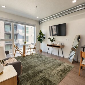 A modern living room with a flat-screen TV, a round table, two chairs, a sofa, a green rug, and a large mirror.