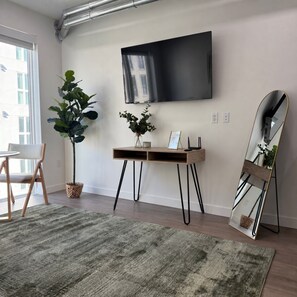 Ein modernes Wohnzimmer mit einem an der Wand befestigten Flachbildfernseher, einem Holztischchen mit einer Topfpflanze, einem Stehspiegel und einem grün gemusterten Teppich.