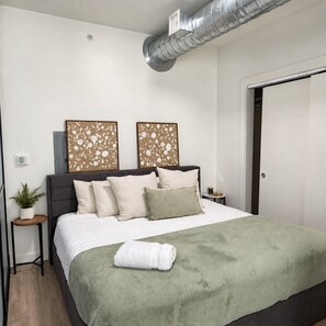 A bedroom with a bed, a bedside table, a potted plant, and a wall-mounted air vent. - Modern Downtown Apt| King Bed+Pool+Prime Location (Salt Lake City)