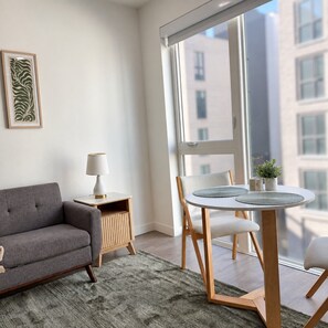 A modern living room with a grey sofa, a small table with a lamp, a round table with chairs, and a large window with a view of buildings. - Modern Downtown Apt| King Bed+Pool+Prime Location (Salt Lake City)
