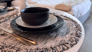 A neatly arranged dining area with a bowl on a plate, a spoon, and a woven placemat. In the background, there is a bed with pillows and a window with blinds.
