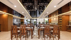 A spacious dining area with wooden tables and chairs, a high ceiling with hanging plants, and a view of the outdoors through large windows. - MAI Luxury Hotel Ninh Binh (Ninh Binh)