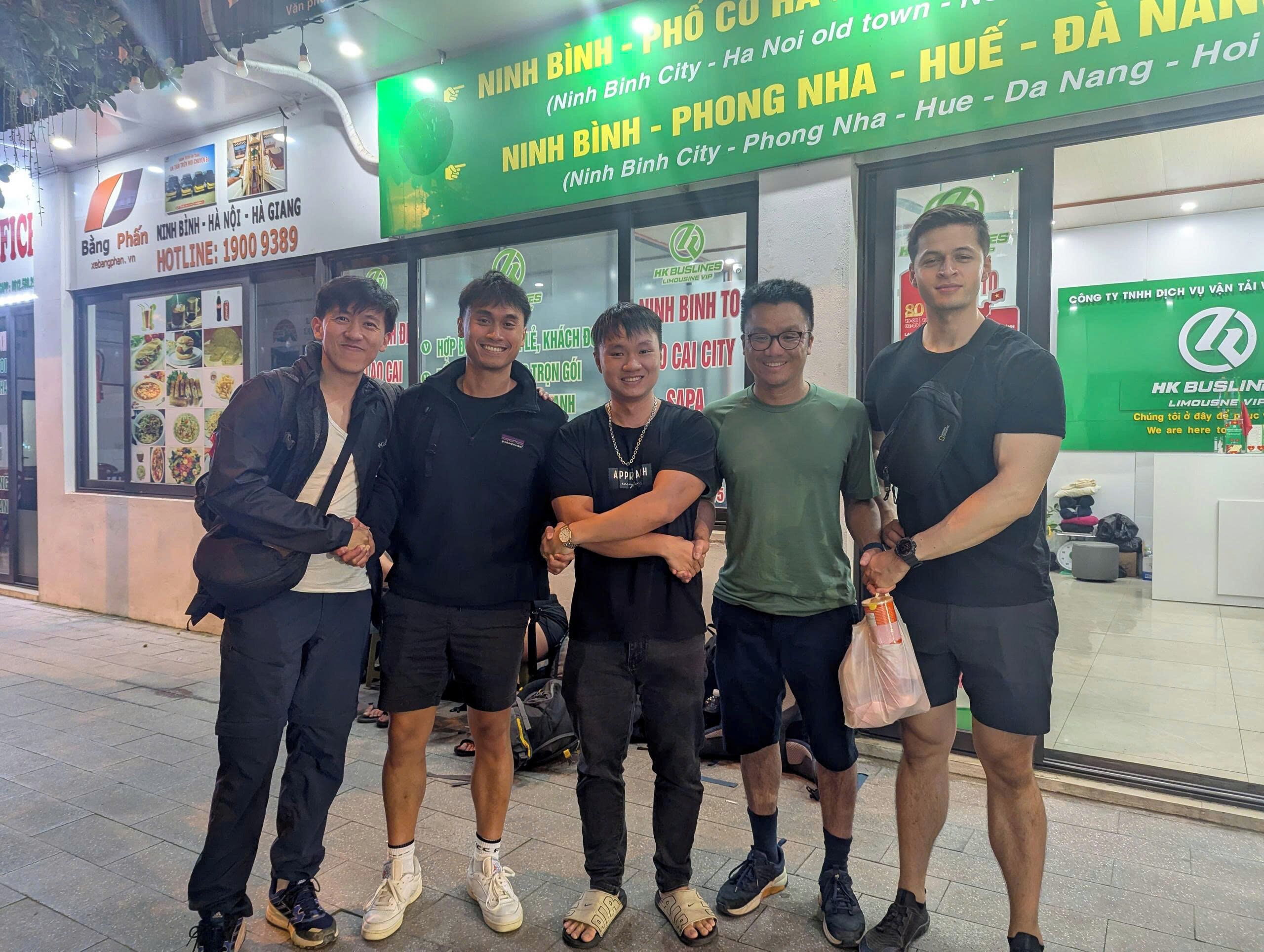 A group of people standing in front of a building with a green signboard listing various locations.