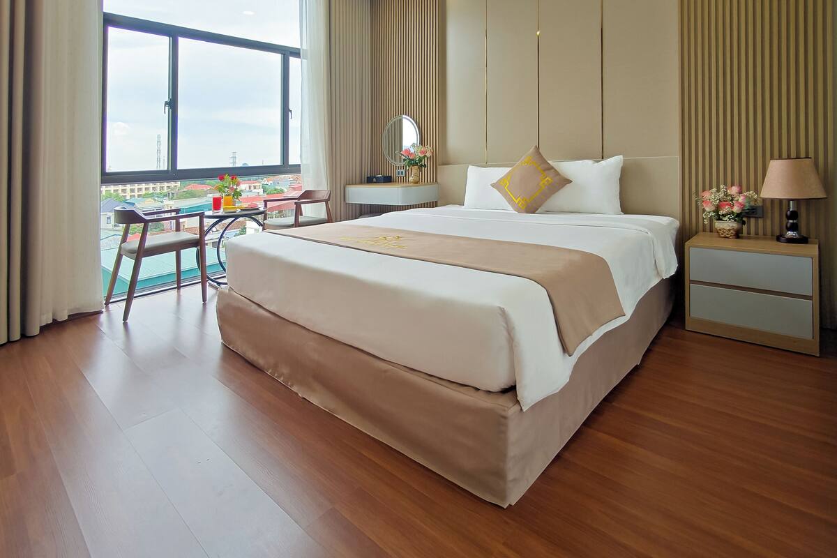 A hotel room with a large bed, a bedside table with a lamp, a chair, and a view of the city through a large window.
