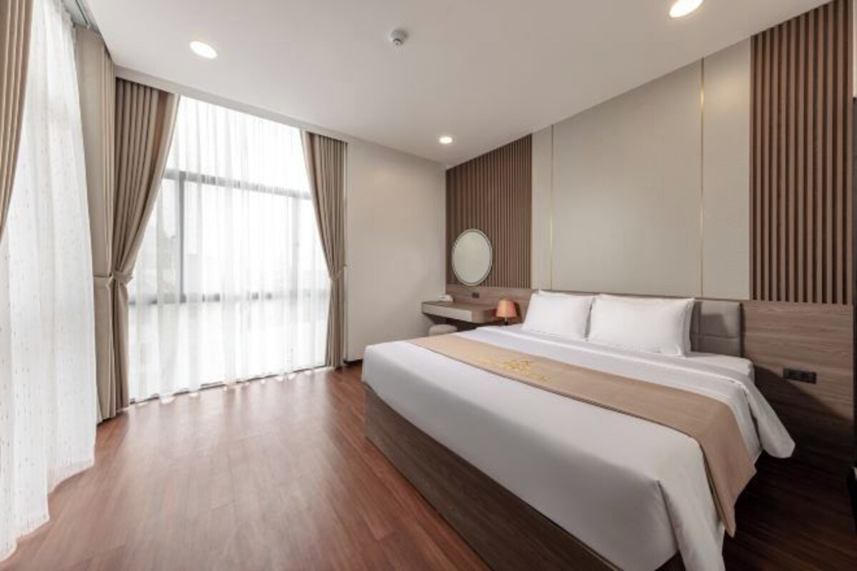 Una habitación de hotel moderna con una cama grande, suelo de madera, una mesita de noche con lámpara y un amplio ventanal con cortinas.