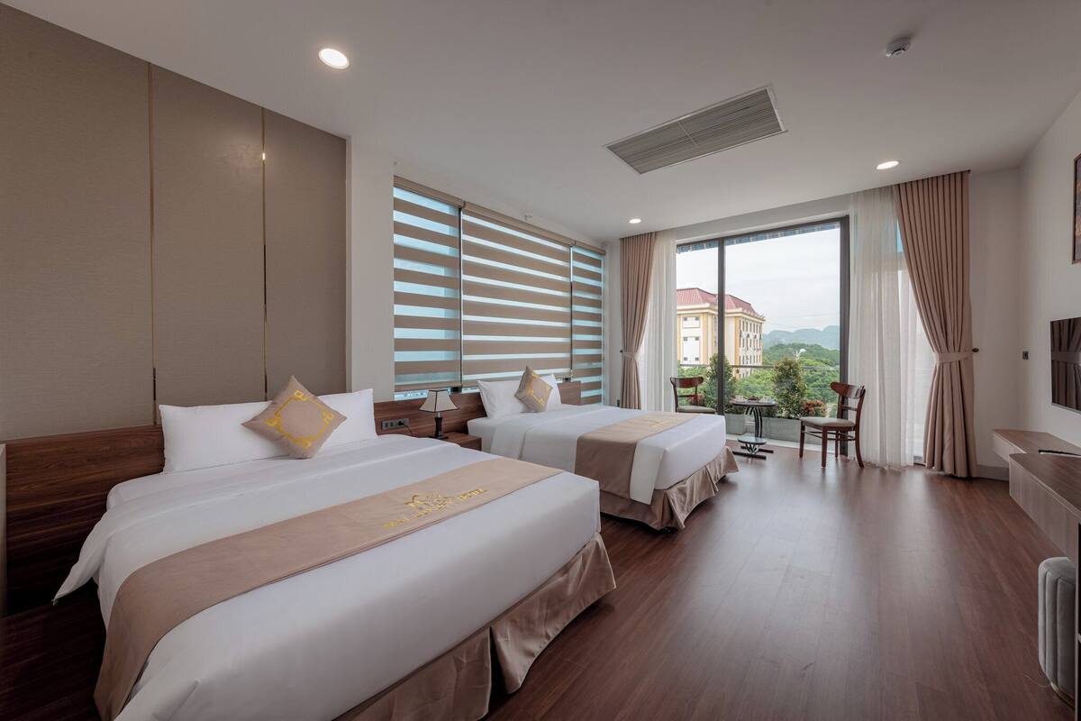 A hotel room with two beds, a large window with blinds, a balcony, and a TV mounted on the wall.
