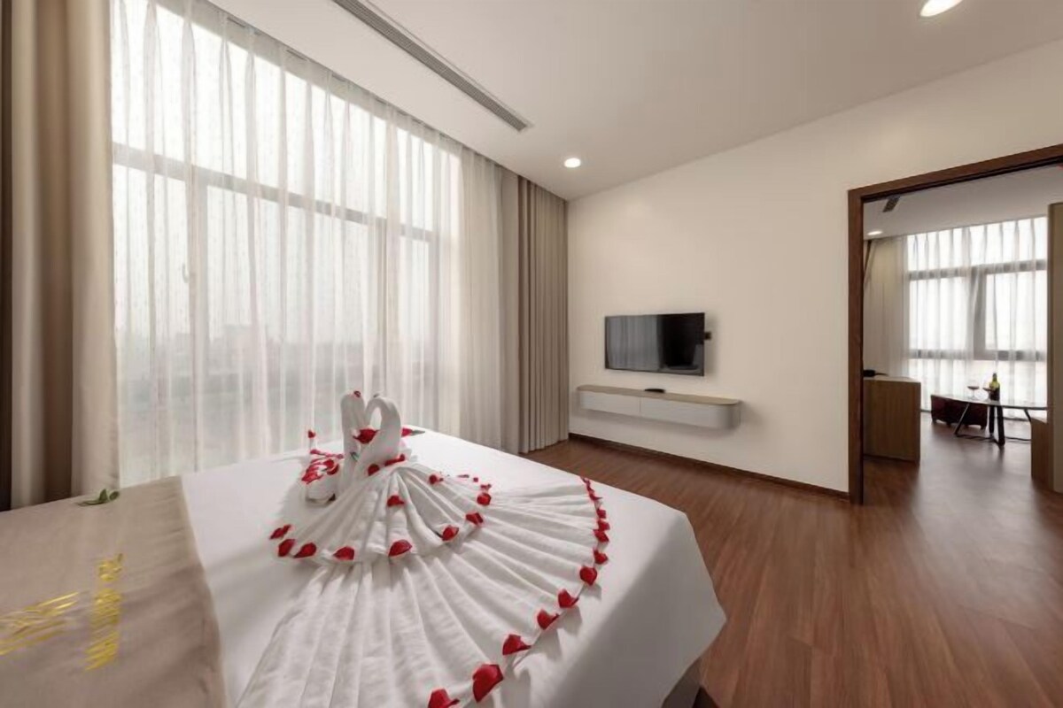 Habitación de hotel con una cama grande, un televisor montado en la pared y vistas a la ciudad desde las ventanas.