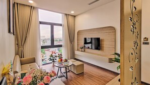 A modern living room with a TV mounted on the wall, a sofa, a coffee table, and a view of the city through large windows. - MAI Luxury Hotel Ninh Binh (Ninh Binh)