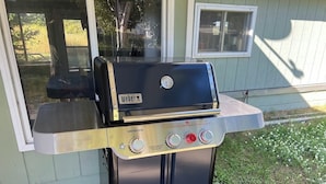 A Weber Genesis portable grill with two burners and a side shelf, positioned on a concrete patio in front of a house. - Agate Beach House - waterfront in Grand Marais (Grand Marais)