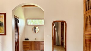 A bathroom with a wooden vanity, a round mirror, and a window with blinds.