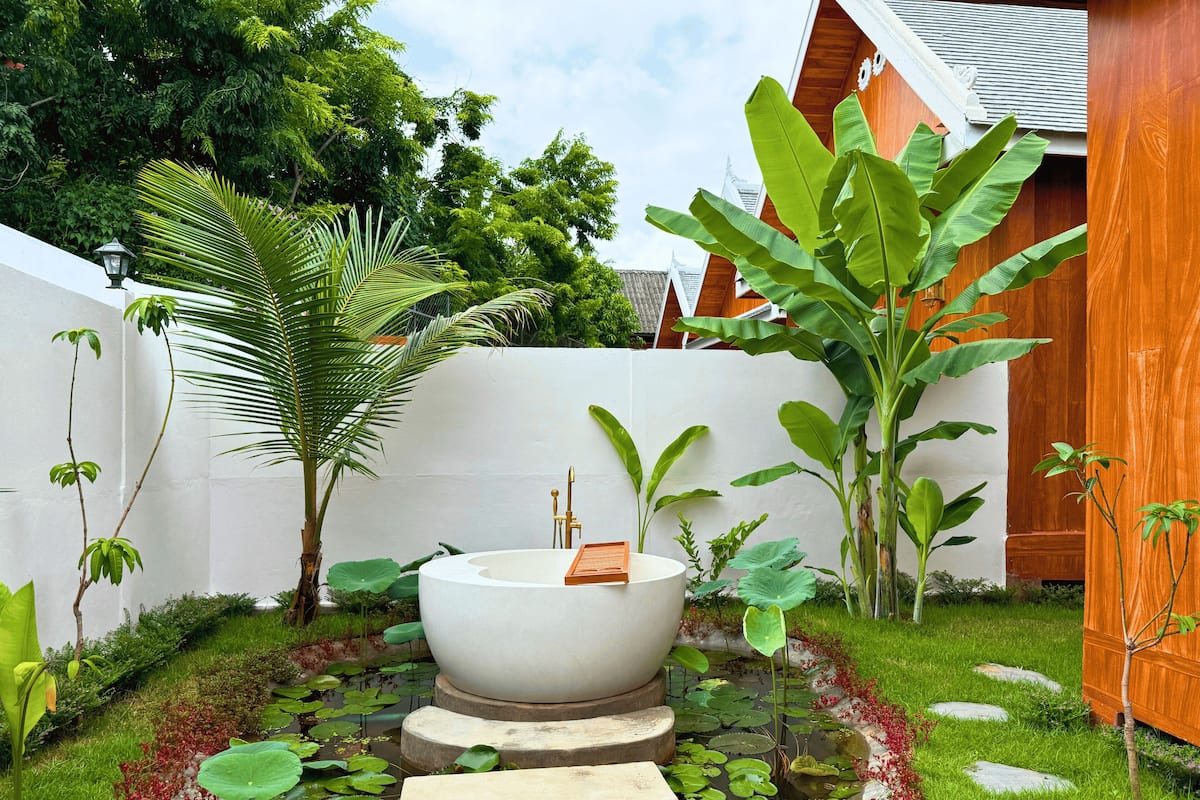 A serene garden with a large white bathtub, lush greenery, and stepping stones leading to a pond with lily pads.