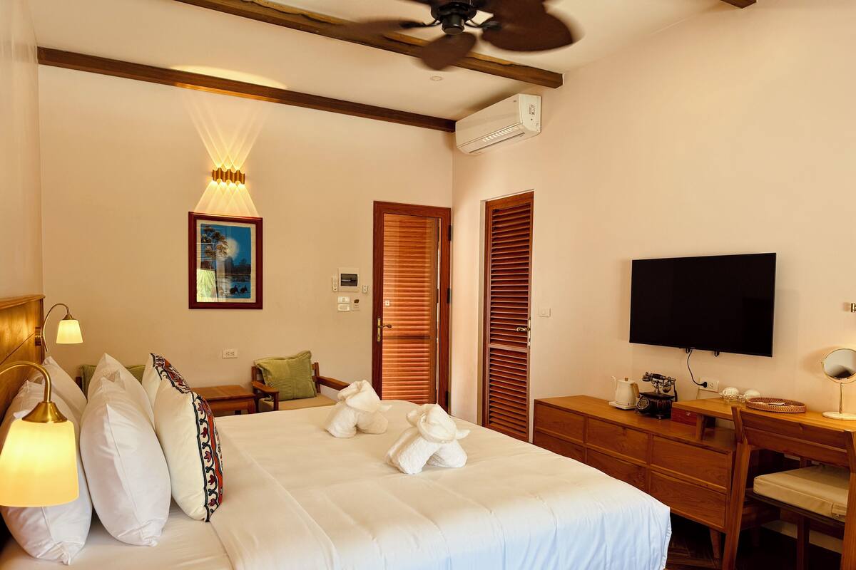 A hotel room with a bed, bedside lamp, a desk with a chair, a television, and a ceiling fan.