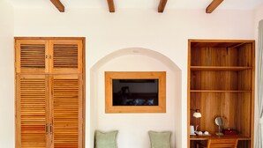 A room with a wooden wardrobe, a bench with cushions, a desk with a lamp, and a wooden bookshelf.