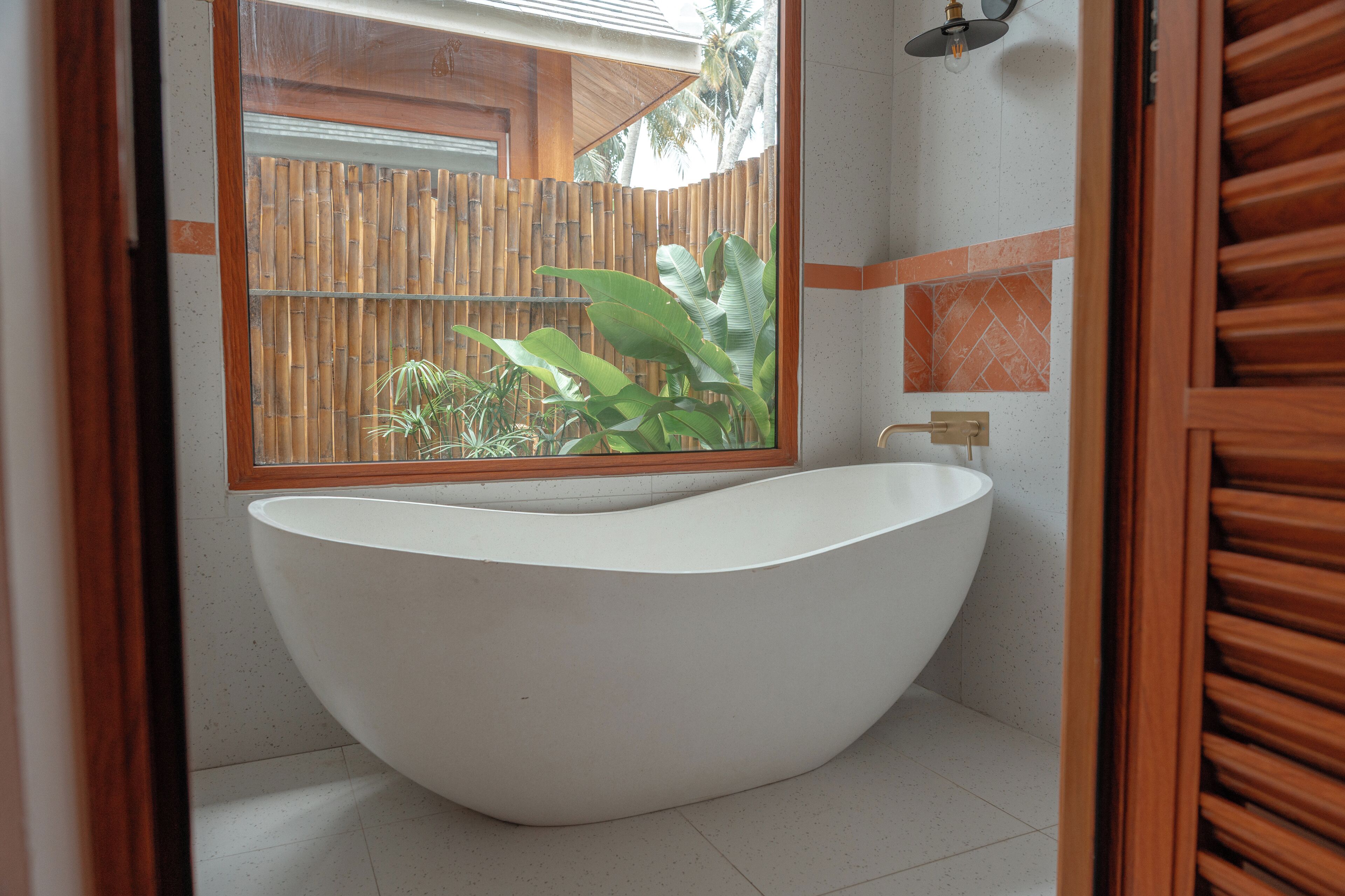 Un bagno moderno con una vasca da bagno indipendente, uno specchio grande e vista su una recinzione di bambù e piante all'esterno.