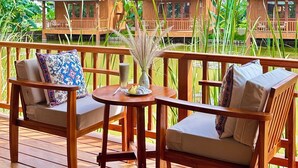 A wooden deck with two sofas and a table, overlooking a tropical resort with wooden bungalows and lush greenery.