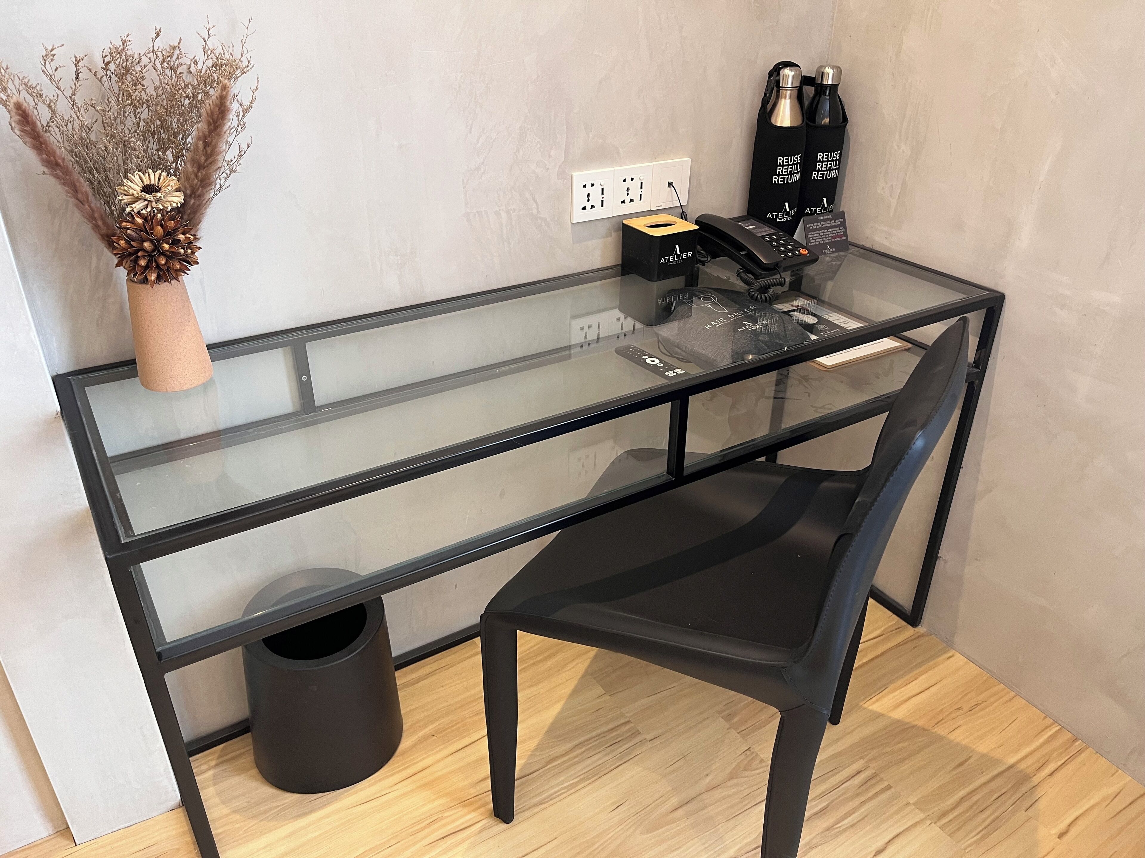 A modern glass desk with a black chair, a vase with dried plants, and a hair care product display.
