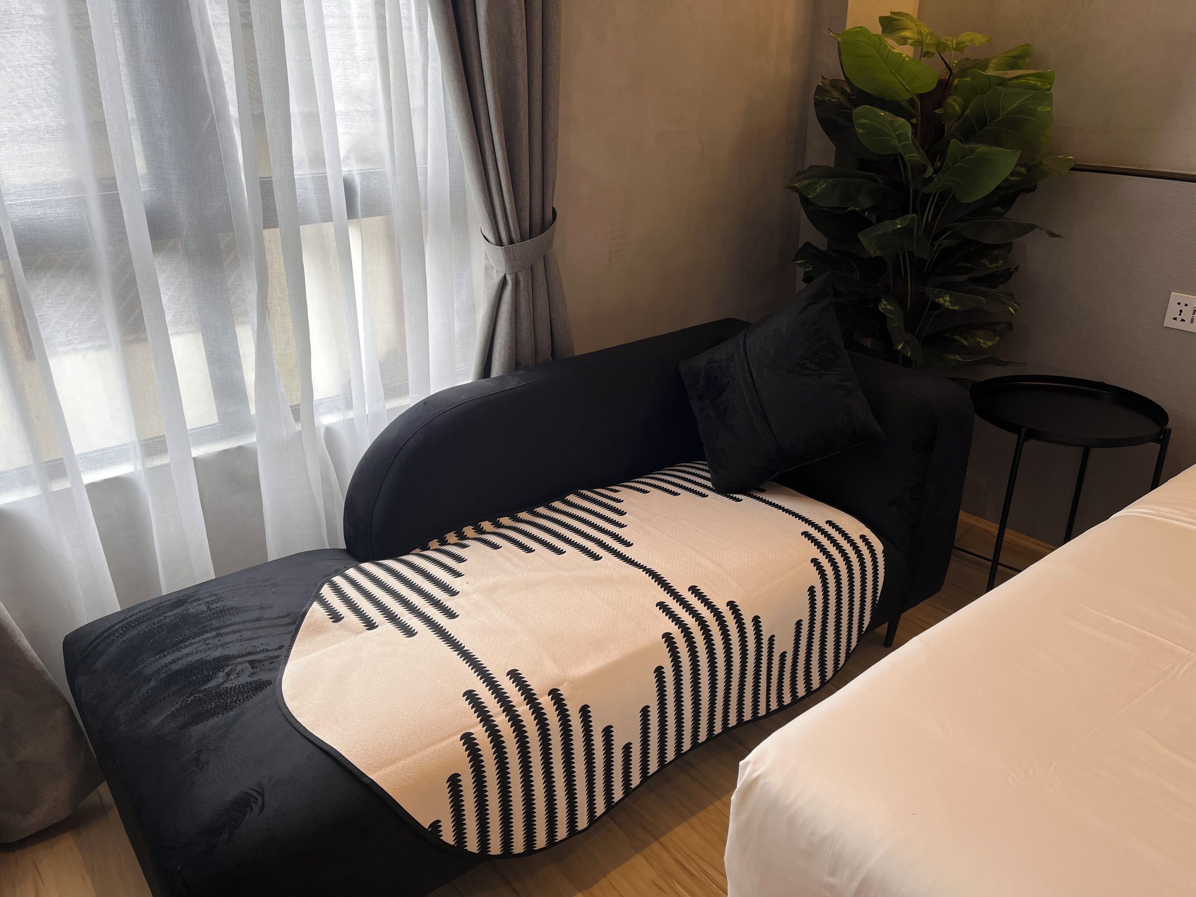 Een hotelkamer met een zwarte bank, een kussen met een wit en zwart patroon, een klein zwart bijzettafeltje, een potplant en een bed met witte beddengoed.