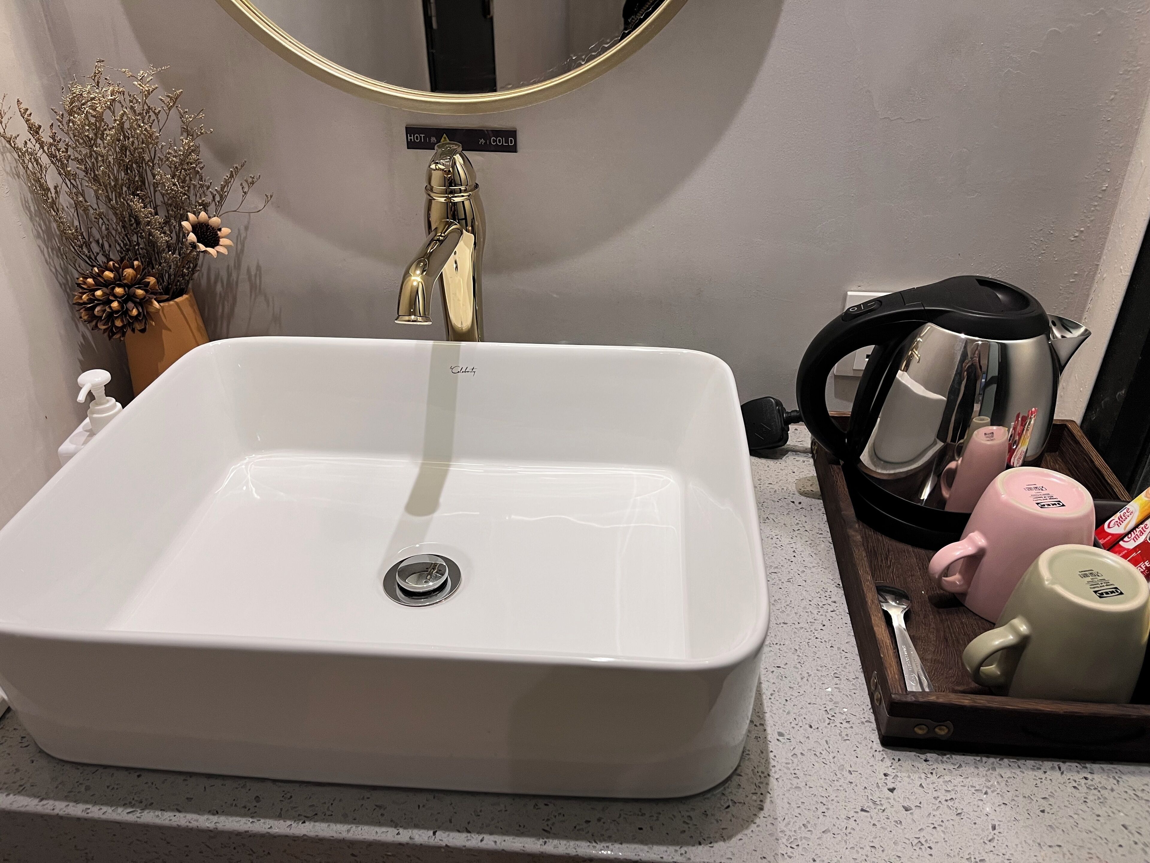 Un baño con un lavamanos blanco, un grifo dorado, un espejo y una bandeja con una tetera, taza y cuchara.
