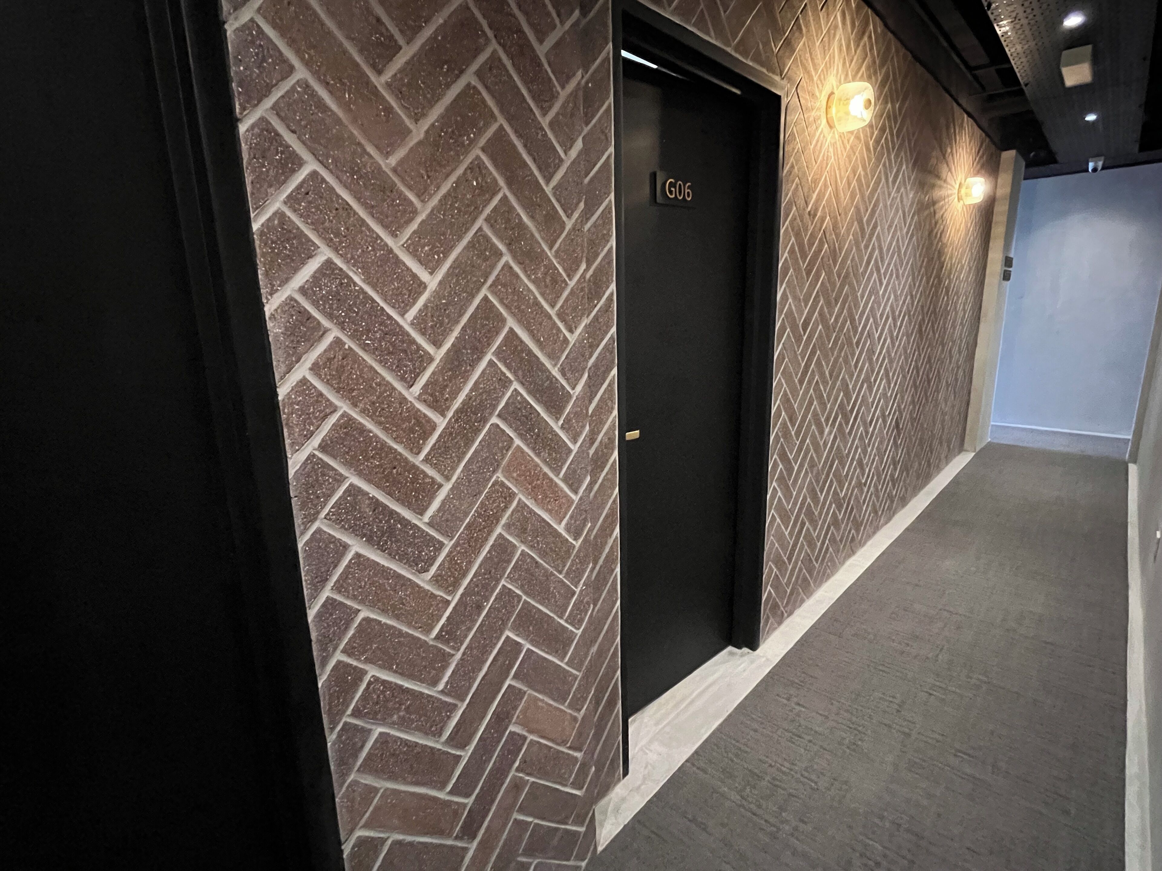 A hallway with a herringbone-patterned brick wall, a black door labeled G06, and a carpeted floor.