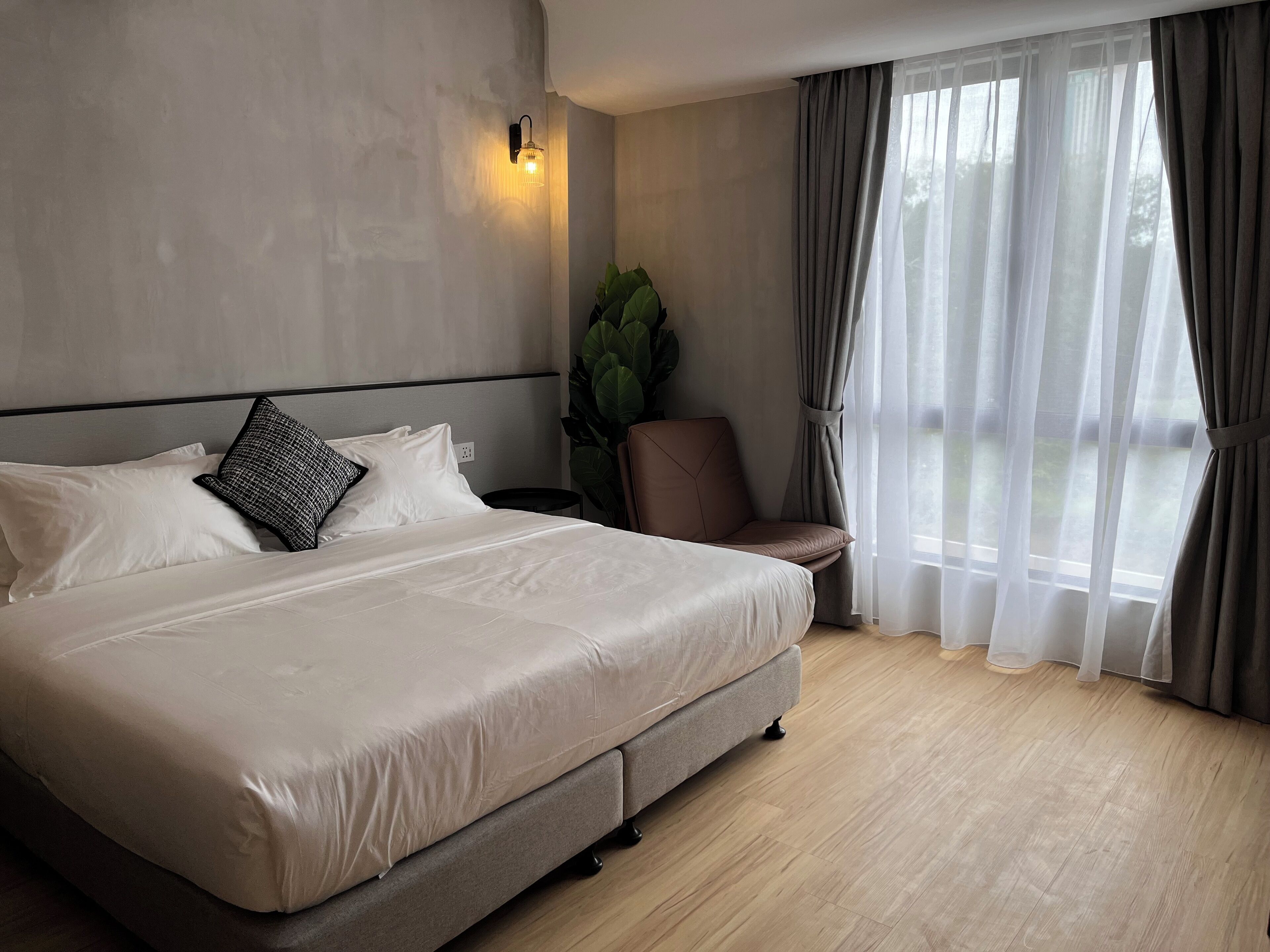 Ein modernes Hotelzimmer mit einem großen Bett, einem Sessel, einer Topfpflanze und Blick durch transparente Vorhänge.