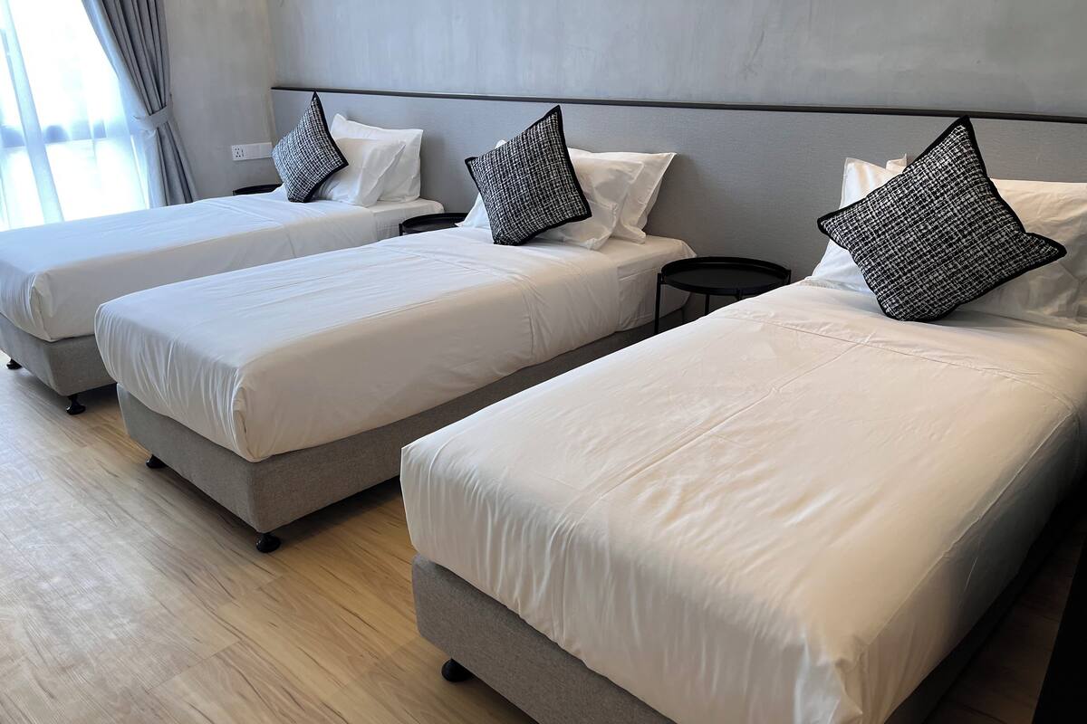 Una habitación de hotel con tres camas individuales, cada una con estructura gris y ropa de cama blanca. Hay una mesita de noche negra pequeña entre las camas.