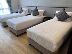Una habitación de hotel con tres camas individuales, cada una con estructura gris y ropa de cama blanca. Hay una mesita de noche negra pequeña entre las camas.