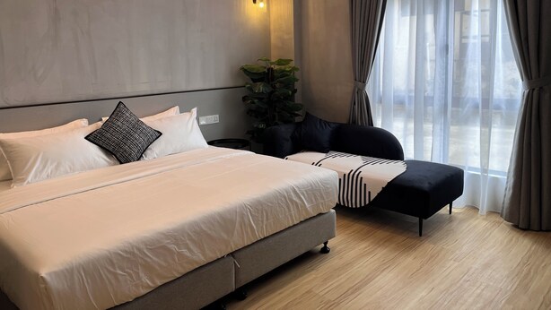 Una habitación de hotel moderna con una cama grande, una mesita de noche, un pequeño rincón de descanso y un amplio ventanal con cortinas.