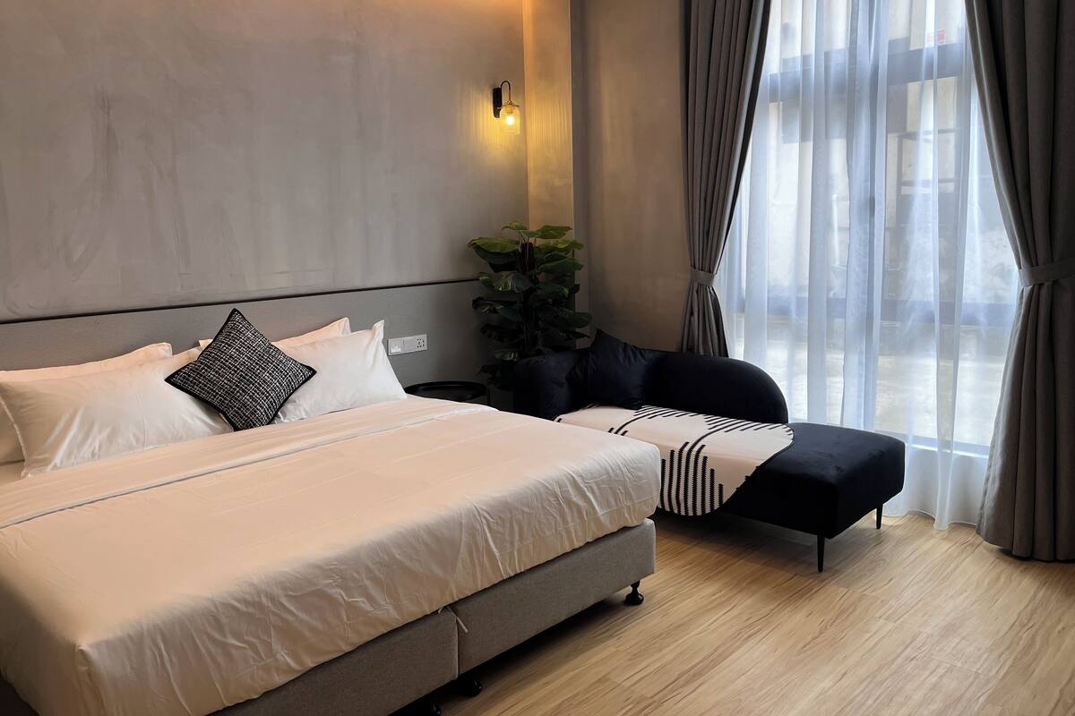 Una habitación de hotel moderna con una cama grande, una mesita de noche, un pequeño rincón de descanso y un amplio ventanal con cortinas.
