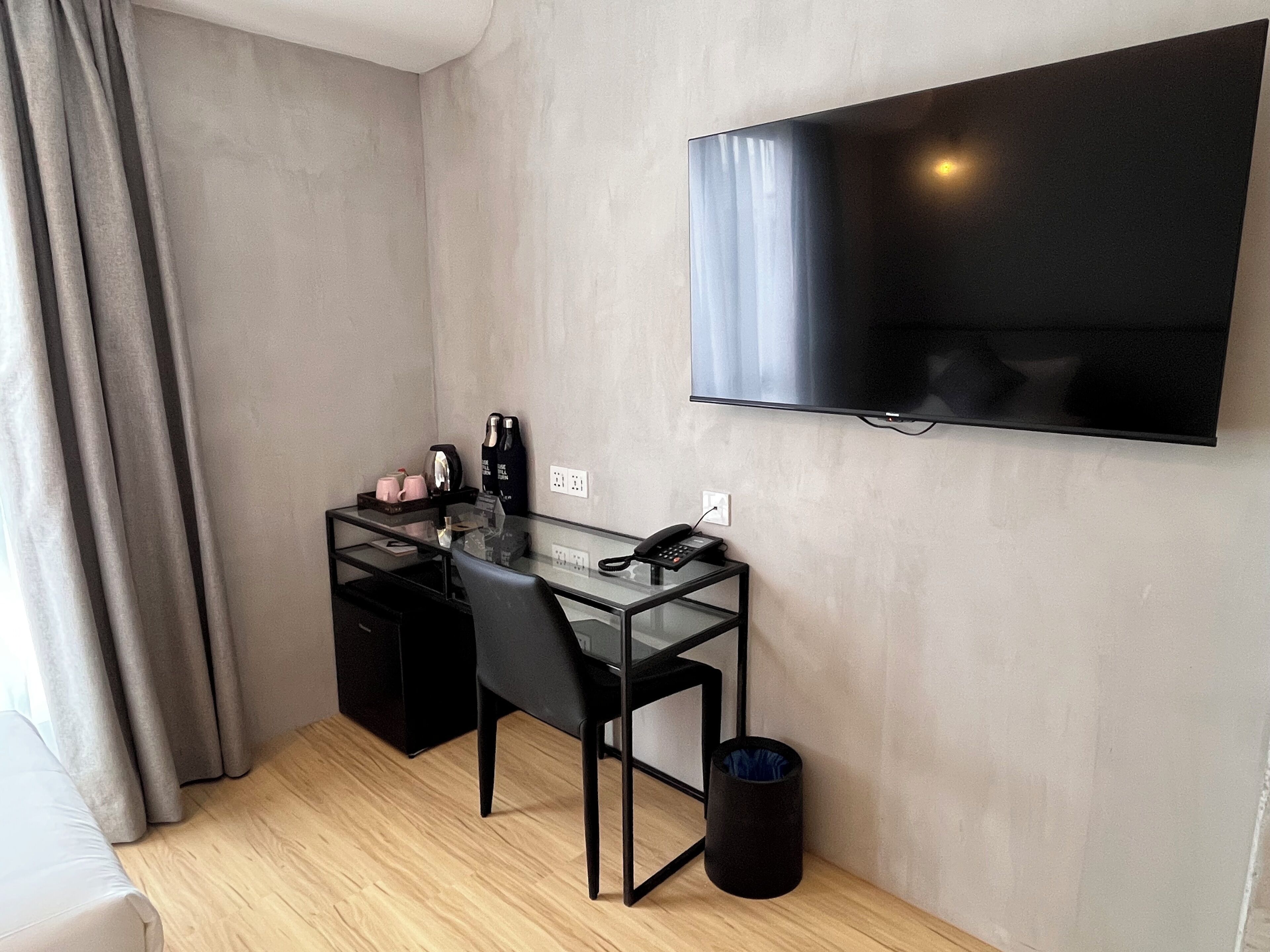 A small, modern room with a glass desk, a black chair, a TV mounted on the wall, and a black trash bin.