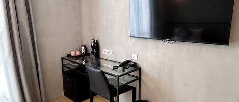 A small, modern room with a glass desk, a black chair, a TV mounted on the wall, and a black trash bin.