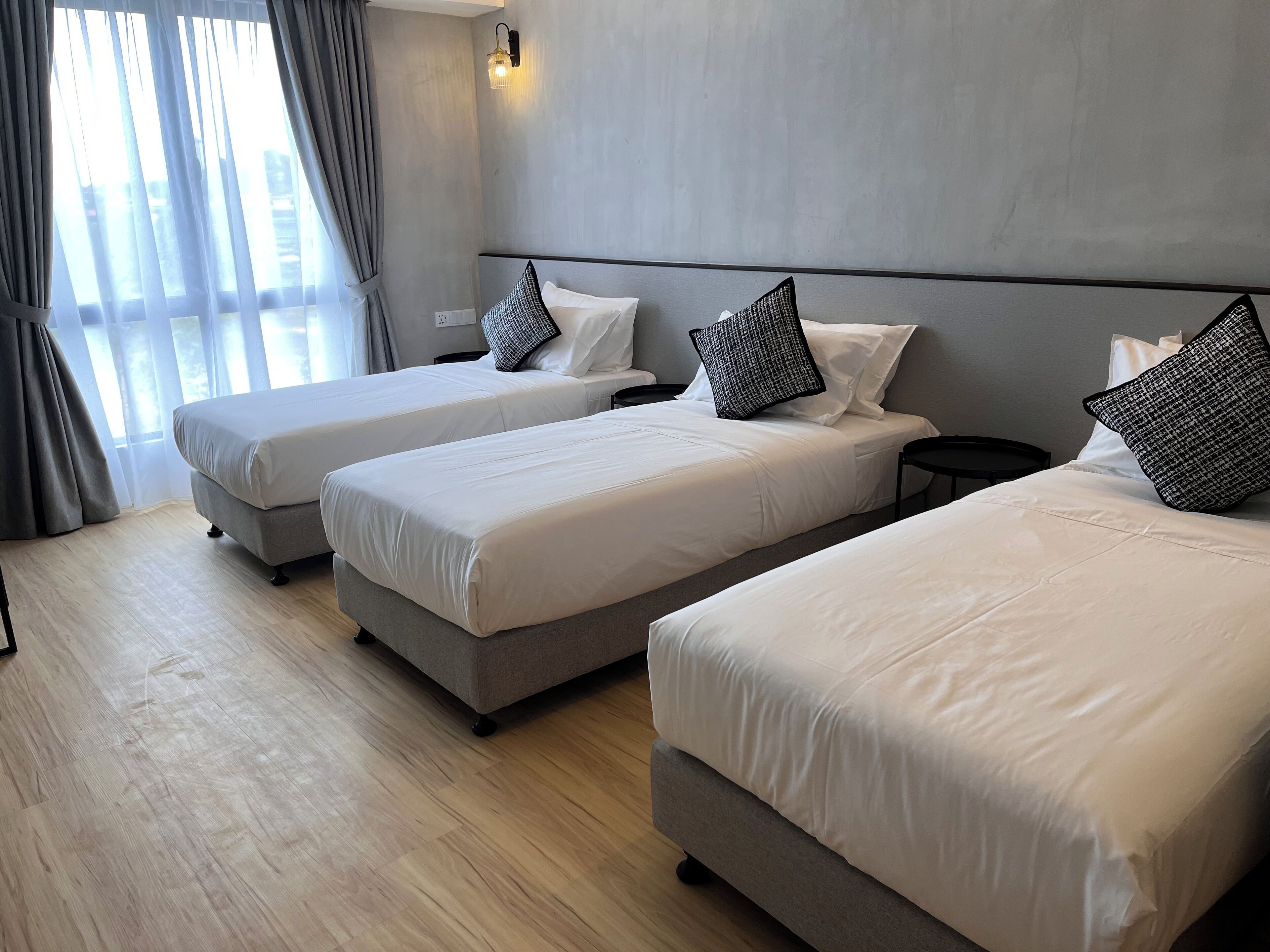 Habitación de hotel con tres camas individuales, una mesita de noche pequeña, una lámpara de pared y una ventana con cortinas.