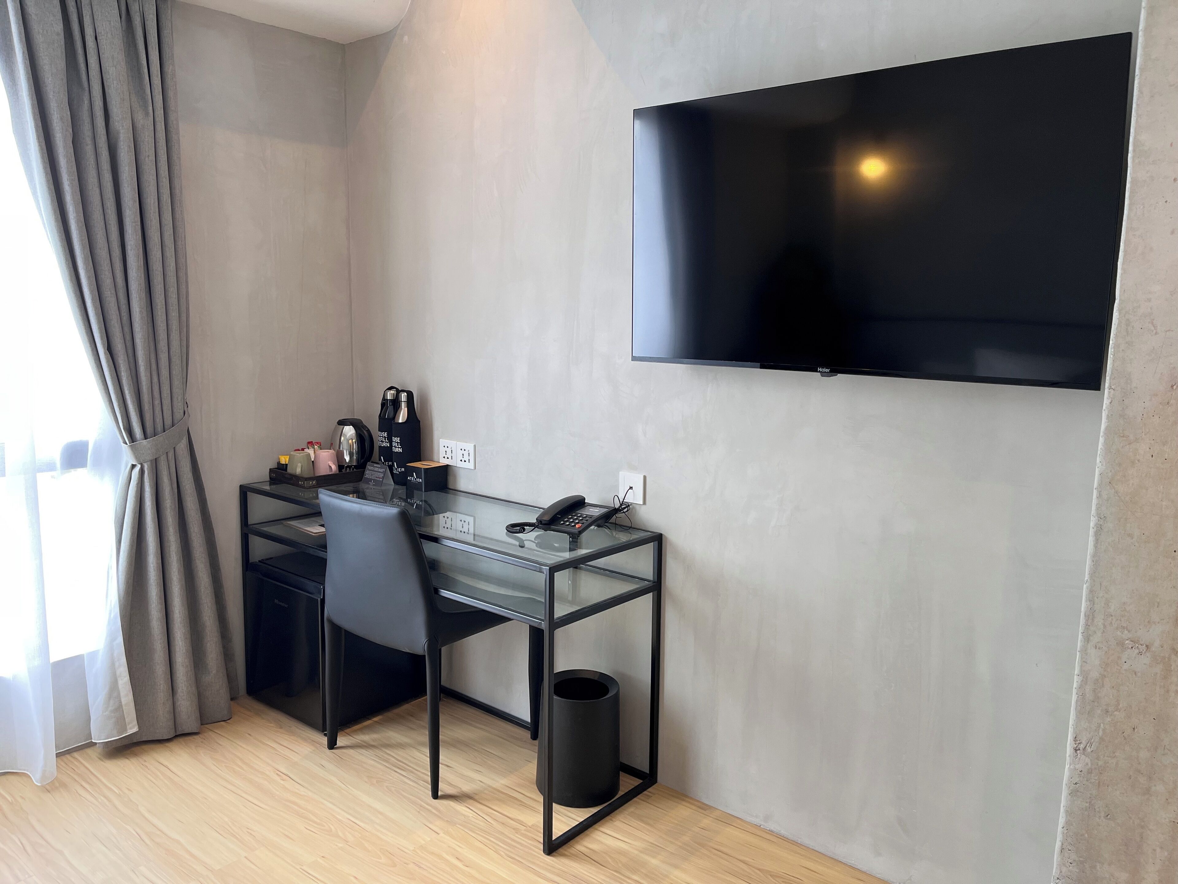 Habitación de hotel moderna con un escritorio de vidrio, una silla negra, un cubo de basura negro, un televisor de pantalla plana montado en la pared y una ventana con cortinas.
